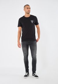 T-shirt en coton noir avec un petit graphisme sur la poitrine gauche, associé à un jean gris foncé déchiré et des baskets noires.
