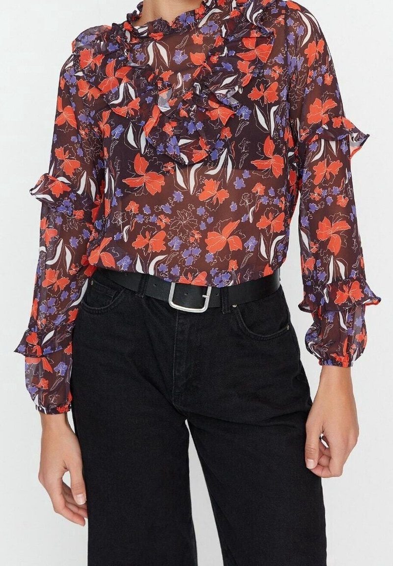 Blouse transparente à manches longues avec un col volanté, présentant un motif floral rouge, violet et blanc sur fond marron foncé. Associée à un pantalon noir.