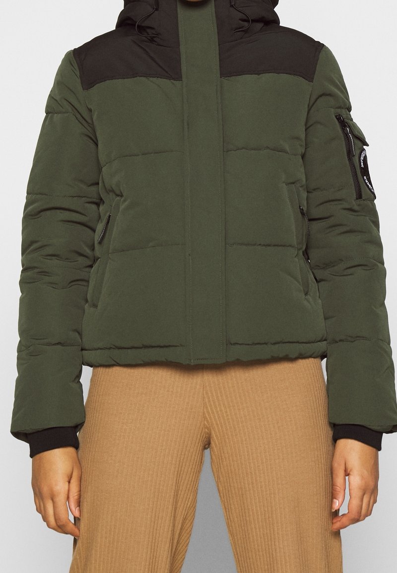 Una persona che indossa un giubbotto imbottito verde e nero e pantaloni a coste beige, in piedi con le braccia rilassate ai lati su uno sfondo neutro.