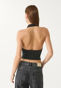 Črn, bleščeč halter crop top z oprijetim dizajnom. Brezhrbtni stil in gladka tekstura dopolnjujeta visoko pasove sive kavbojke.