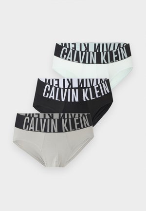 Μυώδης άνδρας με σκούρο δέρμα στέκεται ξυπόλητος φορώντας μαύρα εσώρουχα Calvin Klein, κοιτάζοντας ελαφρώς δεξιά σε απλό φωτεινό φόντο.