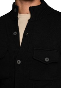 Black knitted cardigan with a high collar, ribbed texture, two front pockets, and button closures. Features gray buttons for fastening.