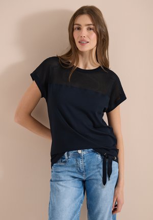 Junge Frau mit einem kurzärmeligen schwarzen Mesh-Oberteil und hellblauen Jeans posiert mit einer Hand in der Hüfte vor einer beigen Wand.