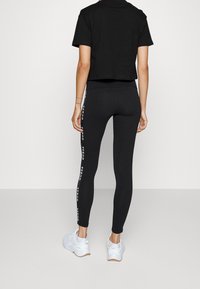 Černé zkrácené tričko, spárované s černými legínami s potiskem loga na straně. Bílé sportovní boty dotvářejí outfit. Hladký textilní povrch.