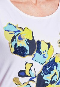 Weißes T-Shirt mit floralem Design in Blau, Gelb und Schwarz. Akzentuiert mit Pailletten und goldenen Nähten entlang der Kanten.