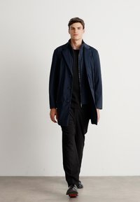 Uomo che cammina in pantaloni neri, camicia nera, cappotto blu navy con bottoni e scarpe nere su uno sfondo bianco uniforme.