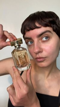 Przezroczysta szklana butelka perfum o geometrycznym kształcie, złota nakrętka, wygrawerowany napis "BOSS" oraz jasno-różowy płyn w środku. Dwie dłonie trzymają butelkę.