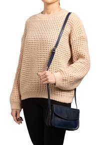 Borsa a tracolla in pelle blu navy con pattina pieghevole, abbinata a un maglione di maglia beige chunky e pantaloni neri attillati. Tracolla regolabile.