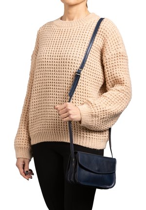 Borsa a tracolla in pelle blu navy con pattina pieghevole, abbinata a un maglione di maglia beige chunky e pantaloni neri attillati. Tracolla regolabile.