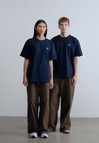 Μπλε βαμβακερά μπλουζάκια συνδυασμένα με χαλαρά καφέ παντελόνια· ένα άτομο έχει ένα τατουάζ ορατό στο χέρι του. Καθημερινά παπούτσια περιλαμβάνουν λευκά και ταμπά παπούτσια.