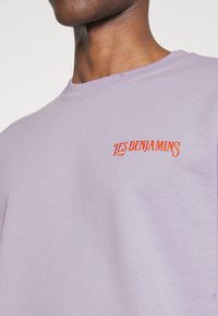 Ingrandimento di una persona che indossa una maglietta viola chiaro con il testo arancione "Les Benjamins" sul petto, collo e linea della mascella parzialmente visibili.