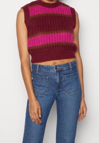 Pull court tricoté à rayures bordeaux, roses et brunes ; design sans manches avec une finition texturée. Associé à un jean bleu taille haute.