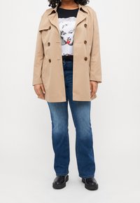 Beżowy trencz z podwójnym zapięciem i dużymi guzikami, noszony na czarnej koszulce z grafiką, w zestawieniu z niebieskimi dżinsami i czarnymi botkami za kostkę.