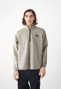 Pullover jacket in beige with a zigzag pattern, quarter-zip front, elasticated cuffs, and logo patch on the left chest. Textured fabric.