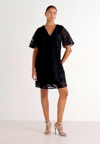 Μαύρο φόρεμα με δαντέλα, που διαθέτει V-λαιμόκοψη και κοντά μανίκια, σε σχήμα Α. Φοριέται με ασημένιες λουρίδες ψηλοτάκουνες γόβες.