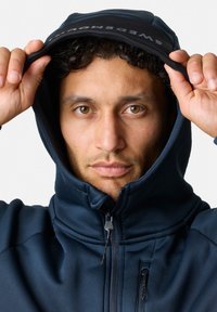 Giacca con cappuccio di colore blu navy, dotata di un colletto alto, chiusura con zip e cappuccio regolabile. Materiale liscio e elastico con dettagli di cucitura ben visibili.