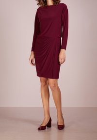 Burgundy knälång klänning med långa ärmar, med drapering vid midjan. Bärs med matchande glansiga klackar. Slät tygstruktur.