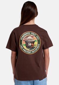 Brun bomull t-shirt med en cirkulär grafisk bild av en björn och texten "Kom ihåg, bara du kan förhindra vildbränder" i grönt och orange.