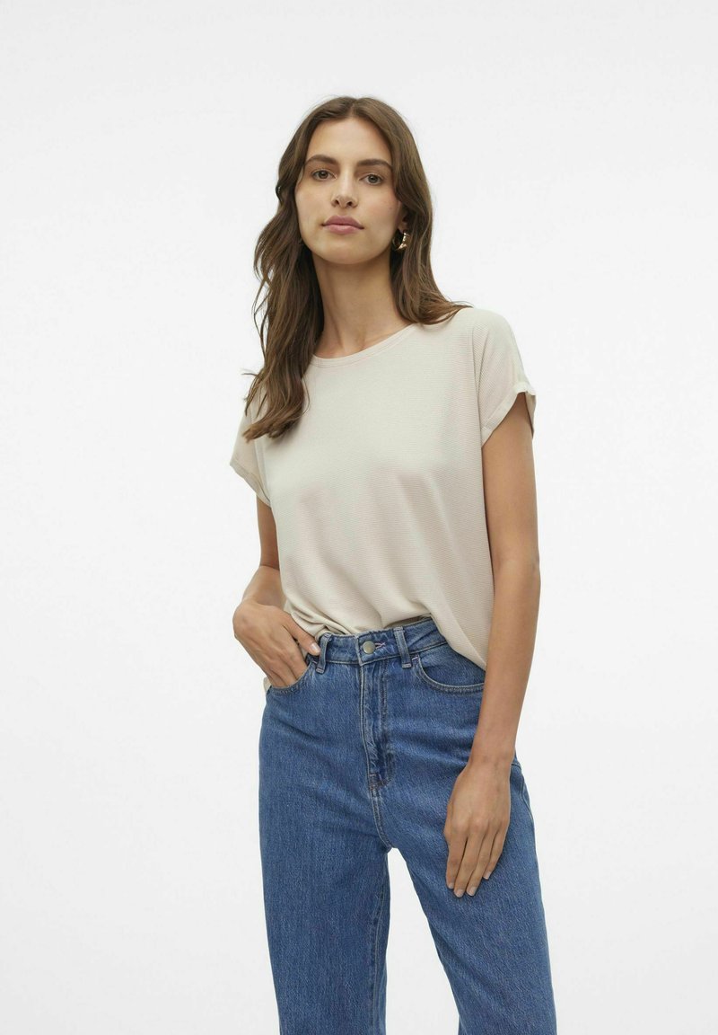 Top corto a maniche corte di un beige chiaro con una vestibilità rilassata, abbinato a jeans in denim blu a vita alta. Tessuto liscio con una leggera trama.