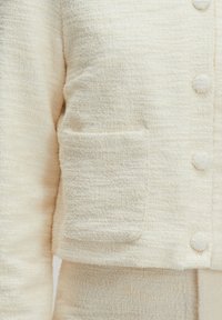 Beige textuur jasje met een korte snit, voorzien van een opgestikte zak en ronde knopen. Het materiaal lijkt zacht en iets donsig.