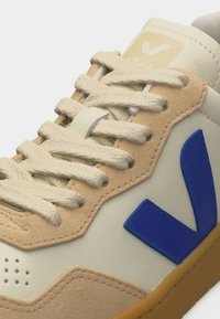 Μπεζ και κρεμ αθλητικό παπούτσι με μπλε λογότυπο "V", κεντημένα κορδόνια, πάνελ από σουέτ και δέρμα, και σόλα από καουτσούκ χρώματος καραμέλας.