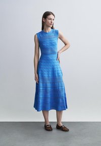 Frau steht vor einer schlichten grauen Wand und trägt ein ärmelloses, blaues, strukturiertes Midikleid sowie Leopardenmuster-Ballerinas, Hand in der Hüfte, schaut nach links.