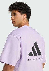Fialové bavlněné tričko s velkým černým logem Adidas a textem "TRAINING" na zadní straně. Krátké rukávy a volný střih.