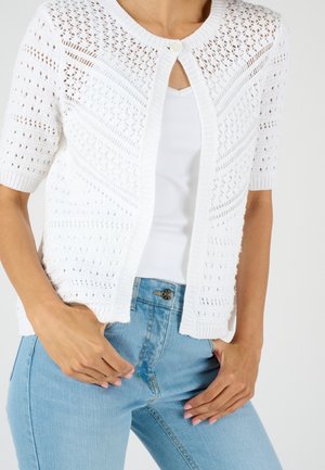 Femme portant un cardigan en maille blanc à manches courtes avec un seul bouton et un jean bleu clair, mains reposant près des poches.