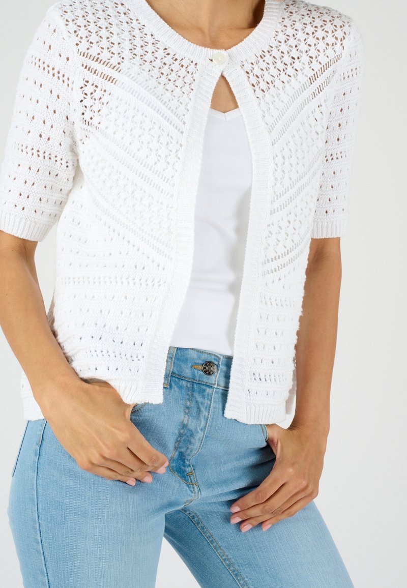 Femme portant un cardigan en maille blanc à manches courtes avec un seul bouton et un jean bleu clair, mains reposant près des poches.