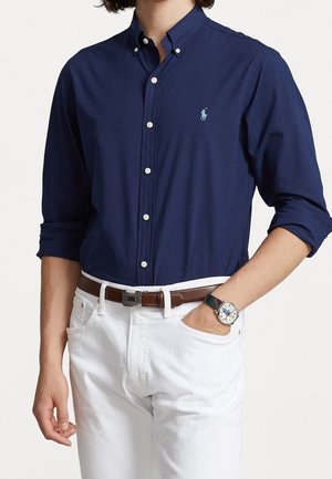 Muž v tmavě modré košili s knoflíky a vyhrnutými rukávy, bílých kalhotách, hnědém pásku a náramkových hodinkách s proužkovaným páskem, ruka v kapse.