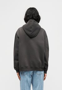 Άτομο με σκούρα μαλλιά μέχρι τους ώμους, που φοράει σκούρο γκρι φούτερ με κουκούλα και ανοιχτό μπλε τζιν με ρίγες, στέκεται με την πλάτη προς το μέρος μας μπροστά σε λευκό φόντο.