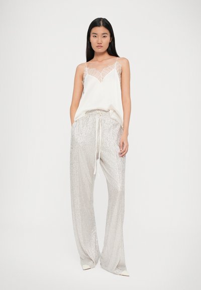 Top alb din dantelă cu bretele subțiri și pantaloni larghi texturați, de culoare argintie, cu talie elastică și șnur. Modelul stă pe un fundal simplu.