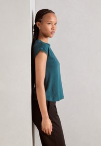 Teal kortermet topp med bølget kant, parret med sorte bukser. Modellen står mot en vegg og viser frem den avslappede passformen og myke teksturen.