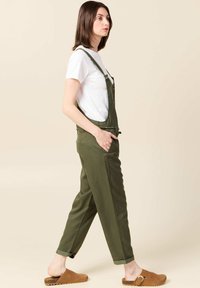 Groene katoenen overalls met verstelbare banden, knoopaccenten en zijzakken, gecombineerd met een wit T-shirt en bruine klompen.