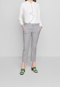 Vit blus med volangdetaljer fram, grå och svart rutiga byxor samt gröna randiga sandaler med klack.