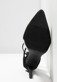Chaussure à talon haut en daim noir avec un bout pointu, dotée d'une semelle lisse et d'un design minimaliste. Légère usure visible sur la semelle.