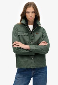 Groene denim jack met een hoge kraag, drukknoopsluiting en twee borstzakken. Heeft een zwart binnenvoering. Gecombineerd met blauwe jeans.