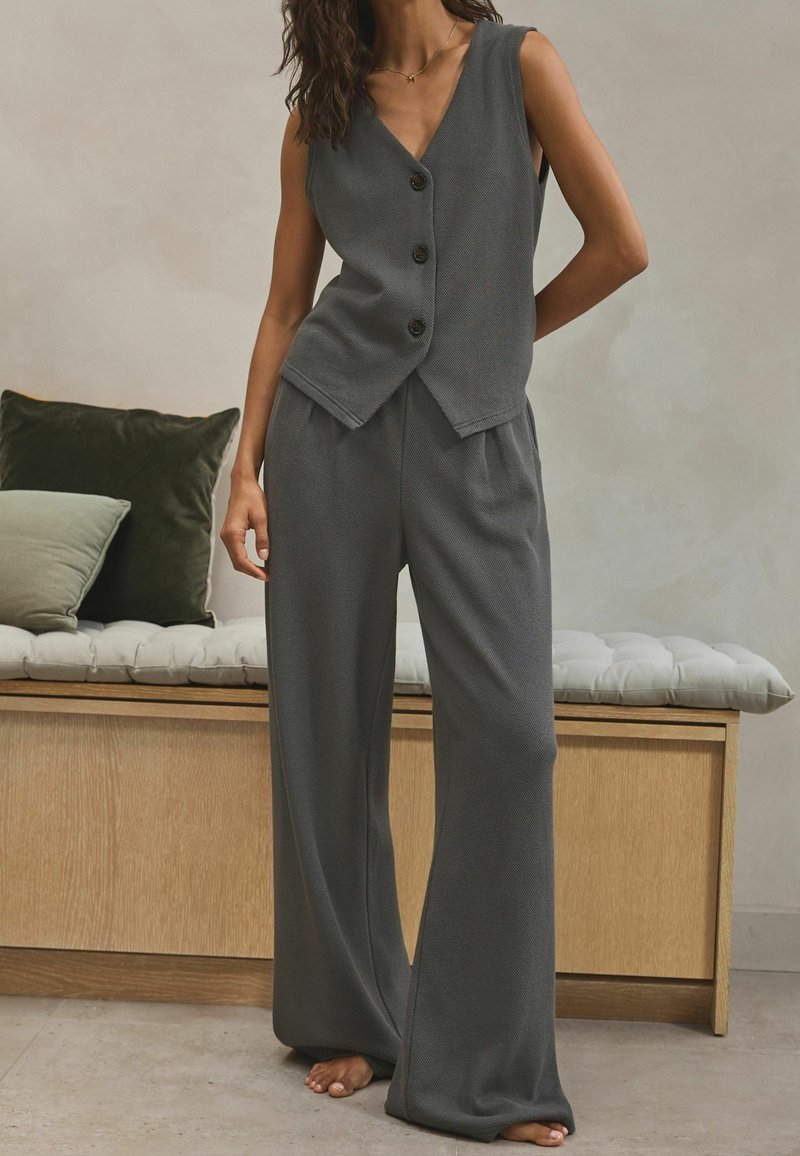 Femme portant un haut sans manches ample gris foncé boutonné et un pantalon à jambes larges, debout pieds nus dans une pièce minimaliste avec un banc en bois et des coussins.