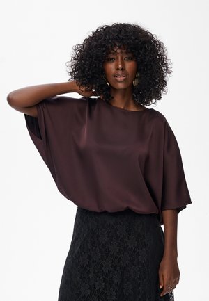 Vrouw met krullend haar in een losse donkerpaarse blouse en een zwarte kanten rok, poserend met één hand achter haar hoofd.