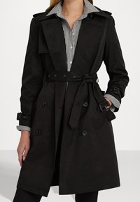 Svart trenchcoat med dubbelknäppt design, midjebälte, axelklaffar och knappade ärmar. Bärs över en randig skjorta.