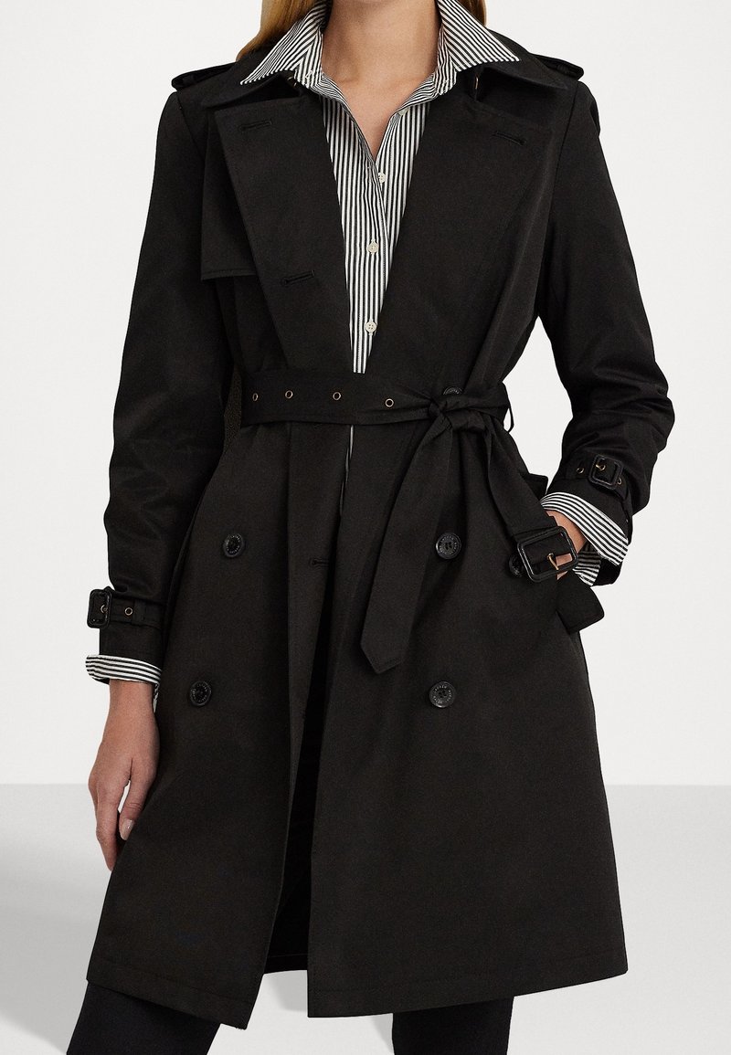 Svart trenchcoat med dubbelknäppt design, midjebälte, axelklaffar och knappade ärmar. Bärs över en randig skjorta.