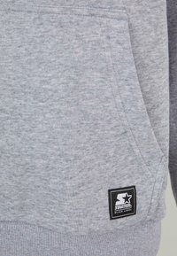 Sweatshirt gris en matériau doux, avec une poche avant et un ourlet côtelé. Une étiquette noire avec un logo d'étoile blanche est visible sur le côté.