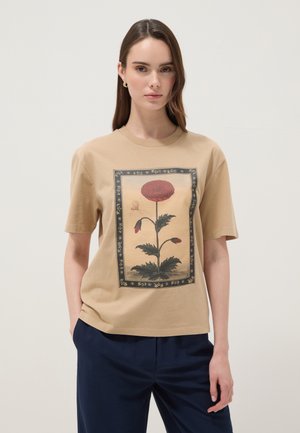 Vrouw die een beige T-shirt draagt met een grote rode bloem en vlinderillustratie, gecombineerd met donkerblauwe broek, staand tegen een effen achtergrond.