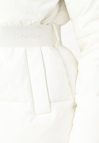 Hvid pufferjakke med detaljer, herunder et struktureret bælte med præget "Calvin Klein" logo, sidelommer og plisserede design elementer.