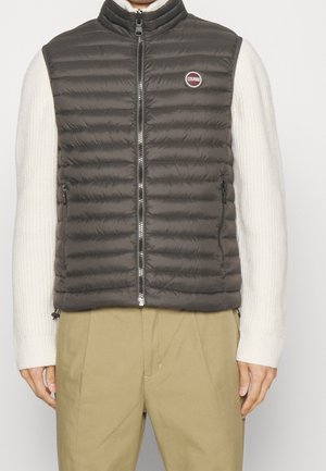 Hombre con chaleco acolchado gris oscuro con cremallera sobre un suéter blanco de punto y pantalones beige, de pie frente a un fondo liso.