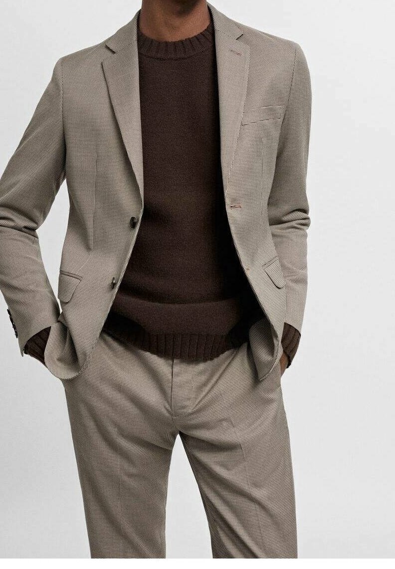 Chaqueta de traje de cuadro gris claro con pantalones a juego, combinada con un suéter marrón de canalé. Presenta solapas con muesca y detalles de botones.