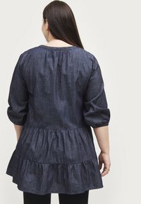 Mørk denim tunika med trekvart lange puffærmer, samlede halsudskæring og flæset kant. Glat tekstur med et lagdelt design.