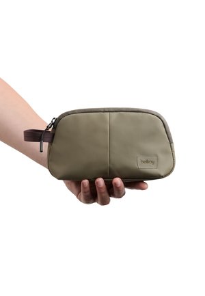Hand hält eine kleine olivgrüne Reißverschlusstasche mit einer braunen Stoffschlaufe und einem "bellroy"-Etikett auf der Vorderseite.