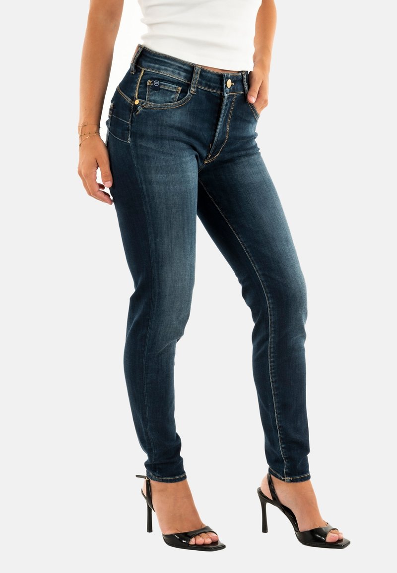 Jean skinny bleu foncé avec une texture lisse, doté d'une fermeture à boutons, de poches latérales et de coutures contrastées. Associé à des sandales à talons noires.
