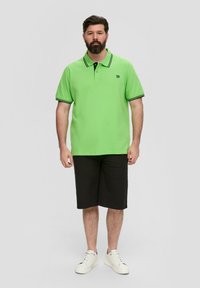 Zielona koszulka polo z czarnymi akcentami, zestawiona z czarnymi szortami i białymi sneakersami. Standardowy krój z kołnierzykiem i zapięciem na dwa guziki.